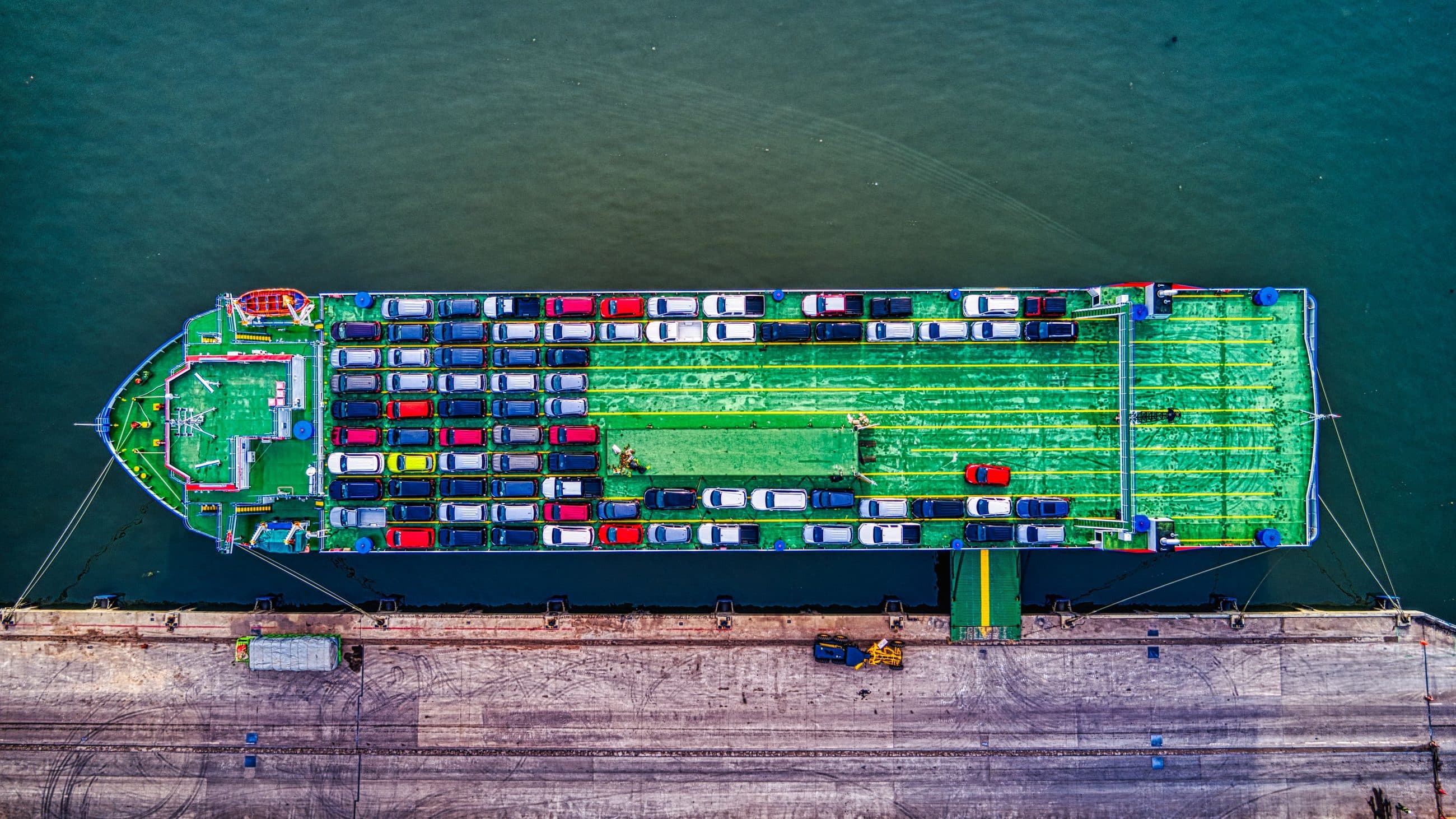 Aerial view of a RoRo vessel at berth, open deck loaded with rows of cars, access ramp lowered