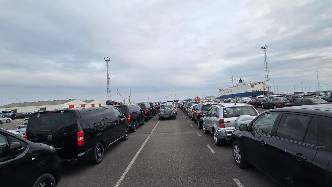 Rows of vehicles lined up at a port terminal, a RoRo vessel moored in the background