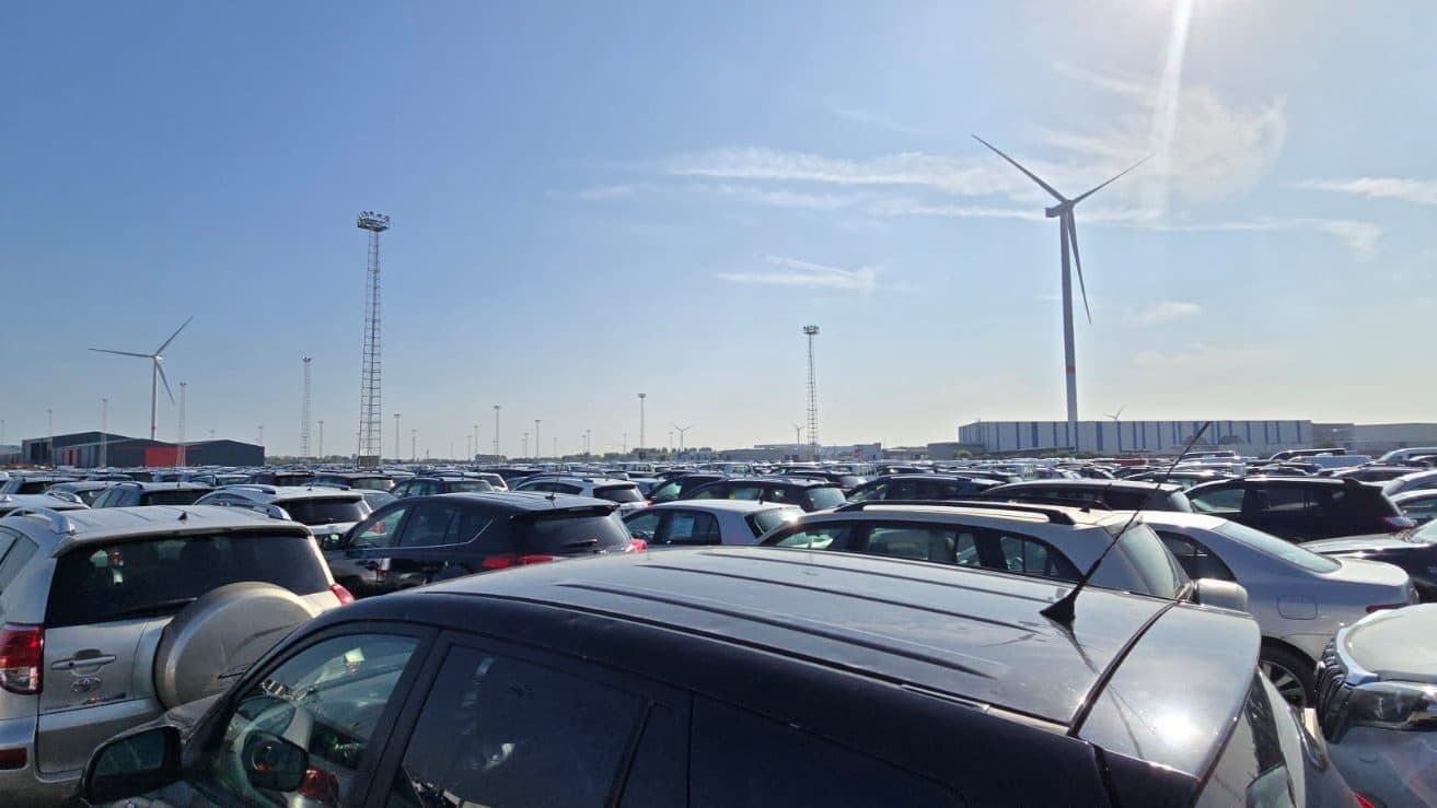 Vast storage yard full of vehicles, wind turbines and warehouses on the horizon